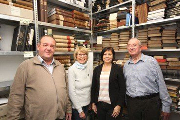 Vier Personen vor Bücherregalen in einem Archiv.